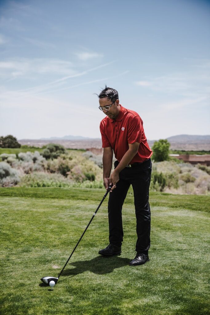 A male golfer in a red shirt prepares to swing on a vibrant golf course under clear skies.