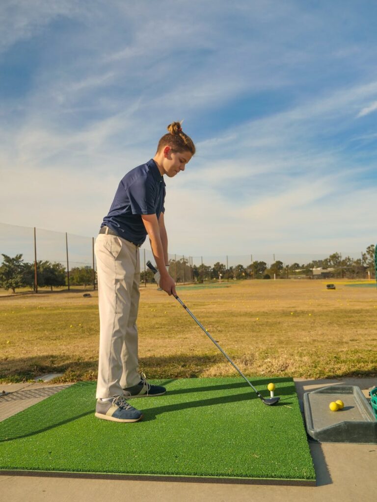 A teenager practicing a golf swing at an outdoor driving range on a sunny day.